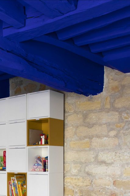 Détail de l'espace dortoir du projet de l'Ecole Montessori Square Paris 18 avec un focus sur le mur en pierres apparentes associé au fameux Bleu Klein très puissant de Ressource Peinture sur le plafond et les poutres structurelles.