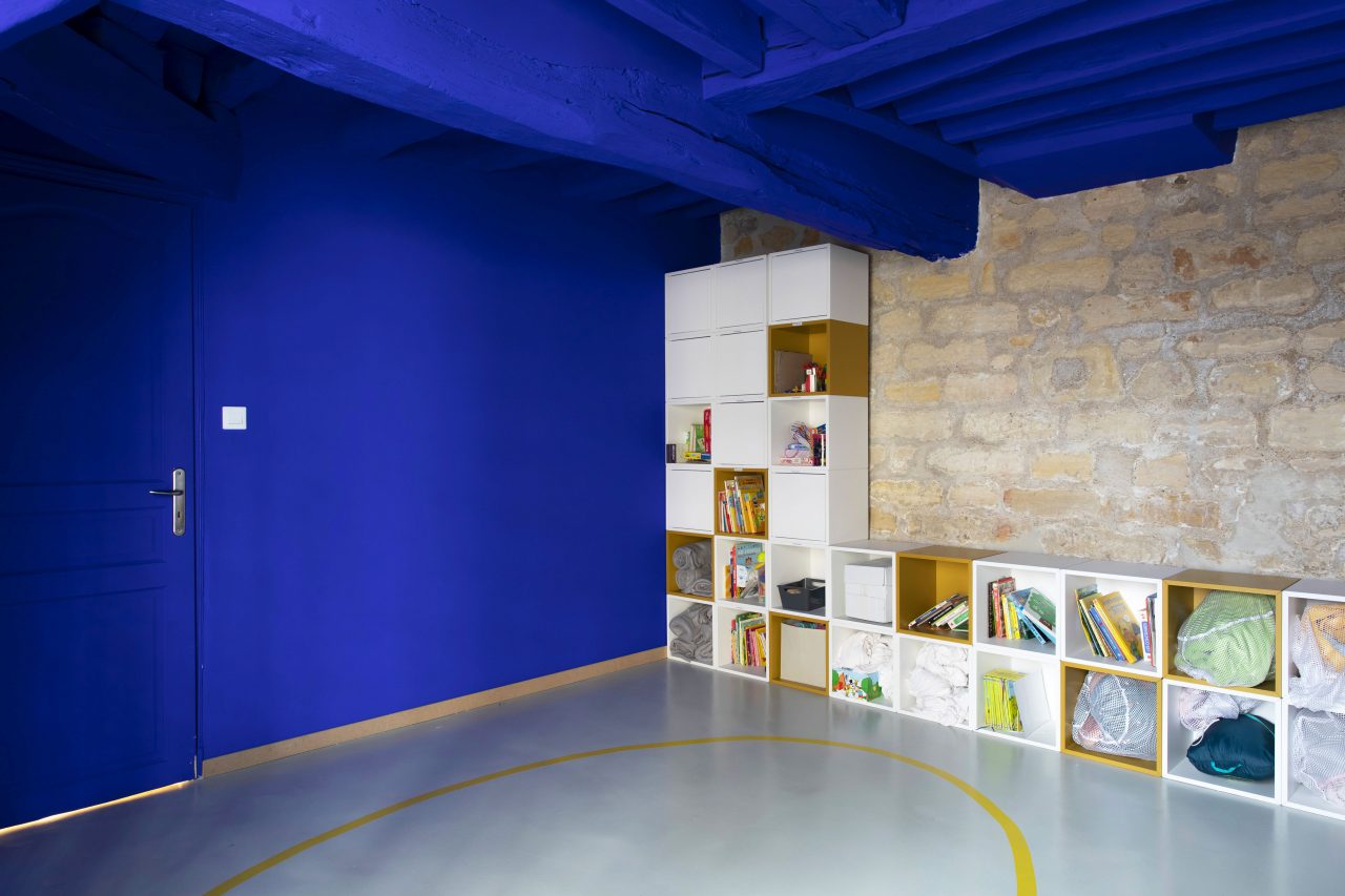 Vue de l'espace dortoir du projet de l'Ecole Montessori Square Paris 18 avec la conservation de son mur en pierres apparentes dynamisé par une mise en peinture des murs adjacents et du plafond avec le fameux bleu Klein de Ressource Peinture.