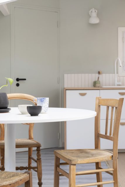 Vue sur l'espace salle à manger et sa jolie table ronde réchauffée de chaises chinées en bois avec en arrière plan la cuisine blanche, bois et vert-de-gris du projet Ribera Paris 16.