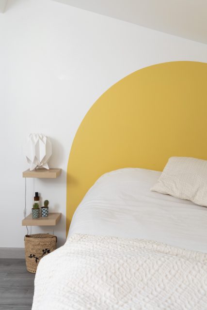 Vue sur la tête de lit graphique en demi cercle peint jaune soleil de cette petite chambre sous les toits de Paris du projet Ribera Paris 16.