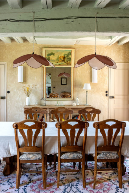 Salle à manger élégante avec un trumeau ancien, une table en bois entourée de chaises sculptées, un tapis Ruggable à motifs Morris & Co, et des suspensions modernes en forme de pétales.