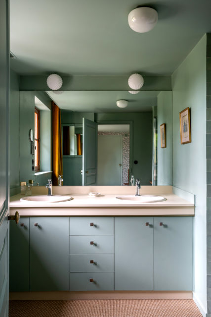 Salle de bain rénovée avec un meuble double vasque vert pastel, robinetterie chromée, miroirs encastrés, éclairage moderne et sol en fibres naturelles, réalisation de rénovation et décoration par Ingrid Marcille dans une maison de campagne en Normandie.