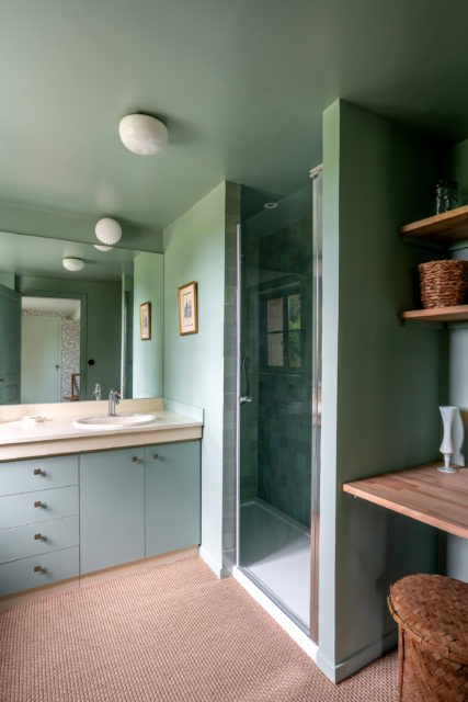 Salle de bain avec douche italienne et meuble double vasque vert pastel, étagères en bois, et sol en fibres naturelles, réalisation de rénovation et décoration par Ingrid Marcille dans une maison secondaire de campagne en Normandie.