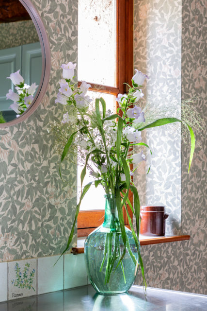 Vase en verre vert avec bouquet de fleurs sauvages placé sur un plan de travail en acier, devant une fenêtre en bois, entouré de papier peint à motifs feuillagés.