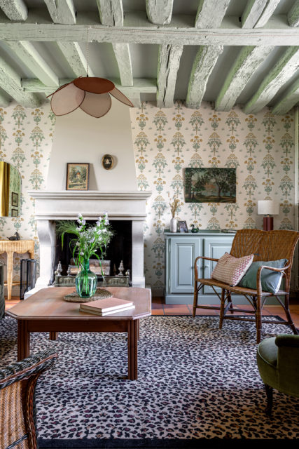 Salon lumineux rénové avec des poutres peintes, un papier peint fleuri, une cheminée en pierre, et un mobilier en osier et bois dans une maison de campagne en Normandie.