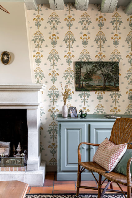 Photo d'un salon de maison de campagne rénové avec une cheminée en pierre, un papier peint floral vintage dans des tons jaunes et verts, un buffet peint en vert pastel, une chaise en rotin avec des coussins, et des accessoires décoratifs élégants. Un design intérieur chaleureux et authentique mêlant charme rustique et raffinement.