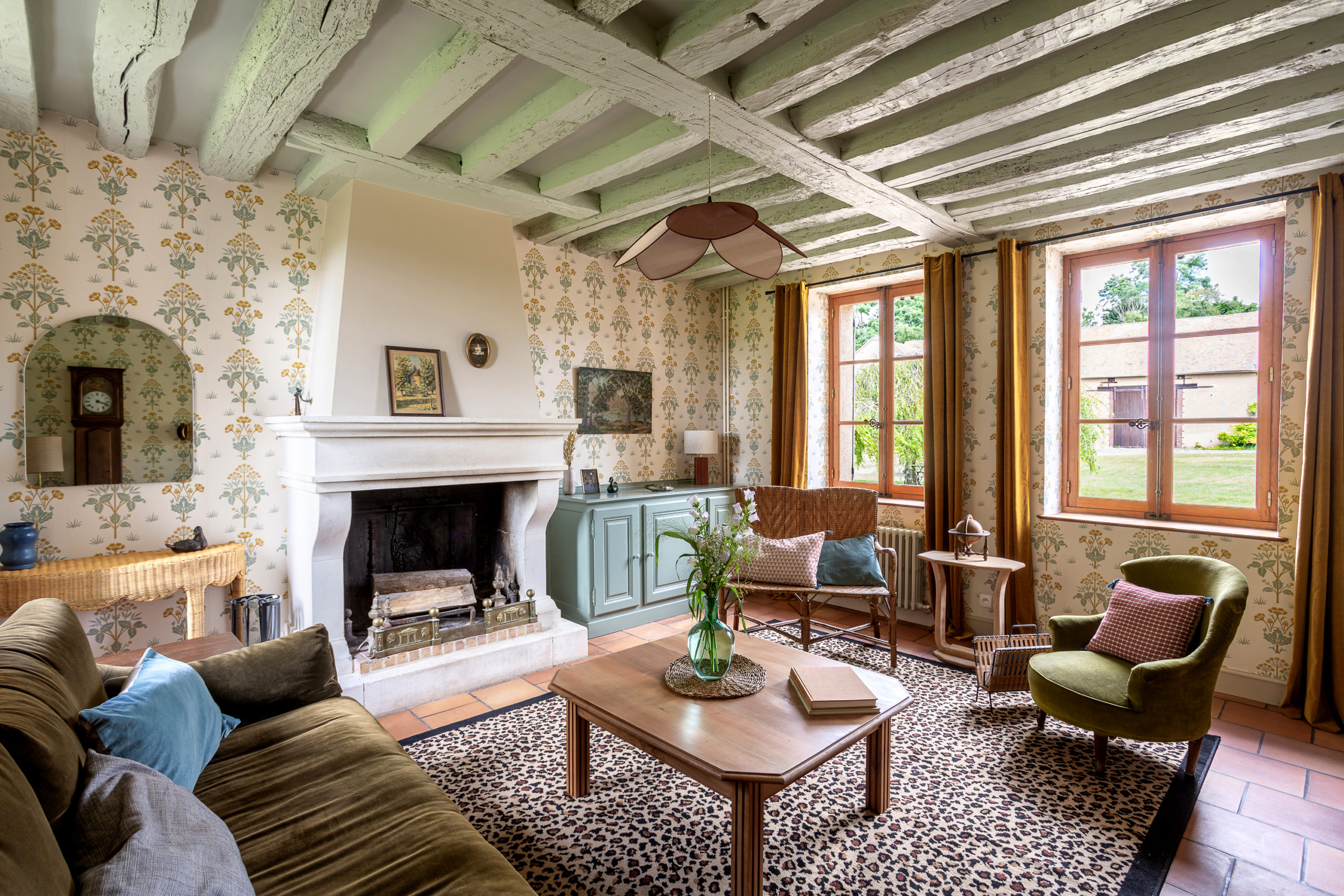 Salon de maison de campagne rénovée avec poutres apparentes, papier peint floral et mobilier en rotin.