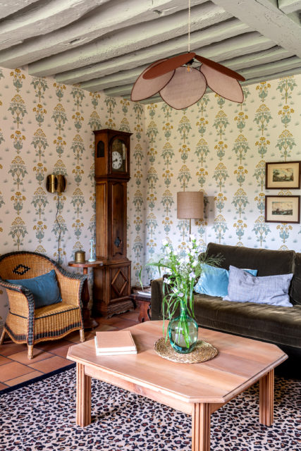 Salon rustique et élégant avec une horloge comtoise en bois, un canapé en velours, une table basse en bois, et des poutres apparentes peintes dans une maison de campagne rénovée en Normandie.