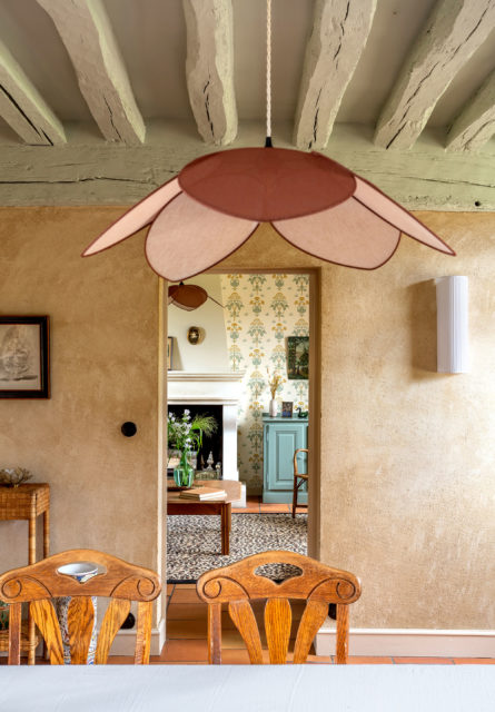 Vue depuis la salle à manger rénovée d'une maison de campagne normande avec des poutres apparentes, une suspension fleur et un intérieur chaleureux aux teintes naturelles.