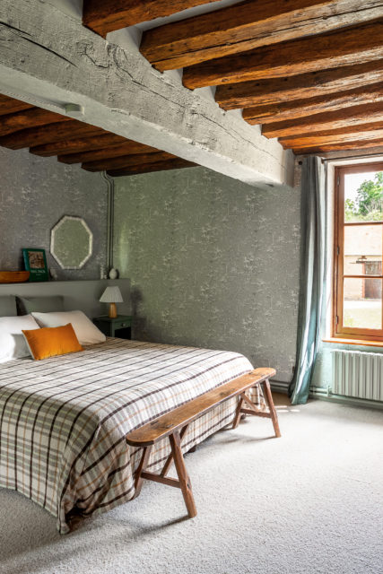 Vue complète de la chambre rénovée d'une maison normande en bord d'eau par Ingrid Marcille, mettant en valeur une ambiance cottage anglais avec un couvre-lit tartan, des poutres apparentes et une tête de lit sur mesure équilibrant les volumes.